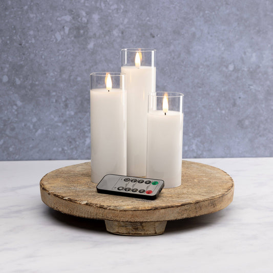 Mellow Monkey set of three white flameless LED pillar candles in glass sleeves with a remote control on a wood tray.