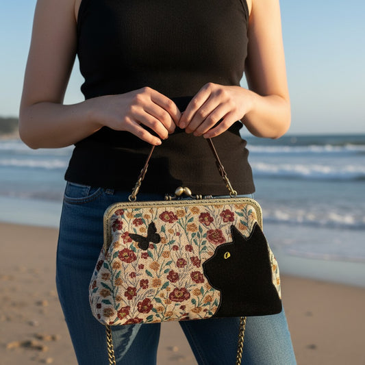 Mellow Monkey floral kisslock bag with black velvet cat and butterfly appliqué held by a person on a beach.