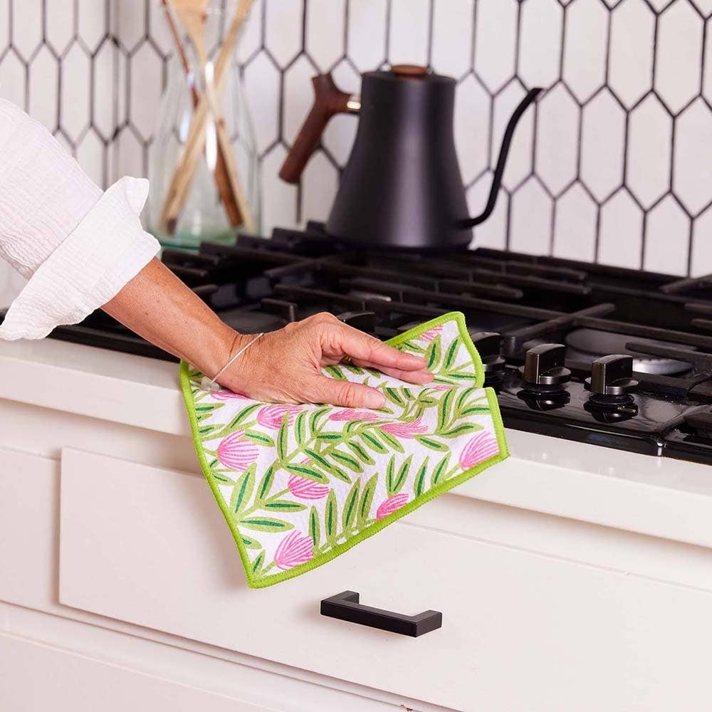Mellow Monkey floral dish towel with pink flowers and green trim being used to wipe a kitchen counter.