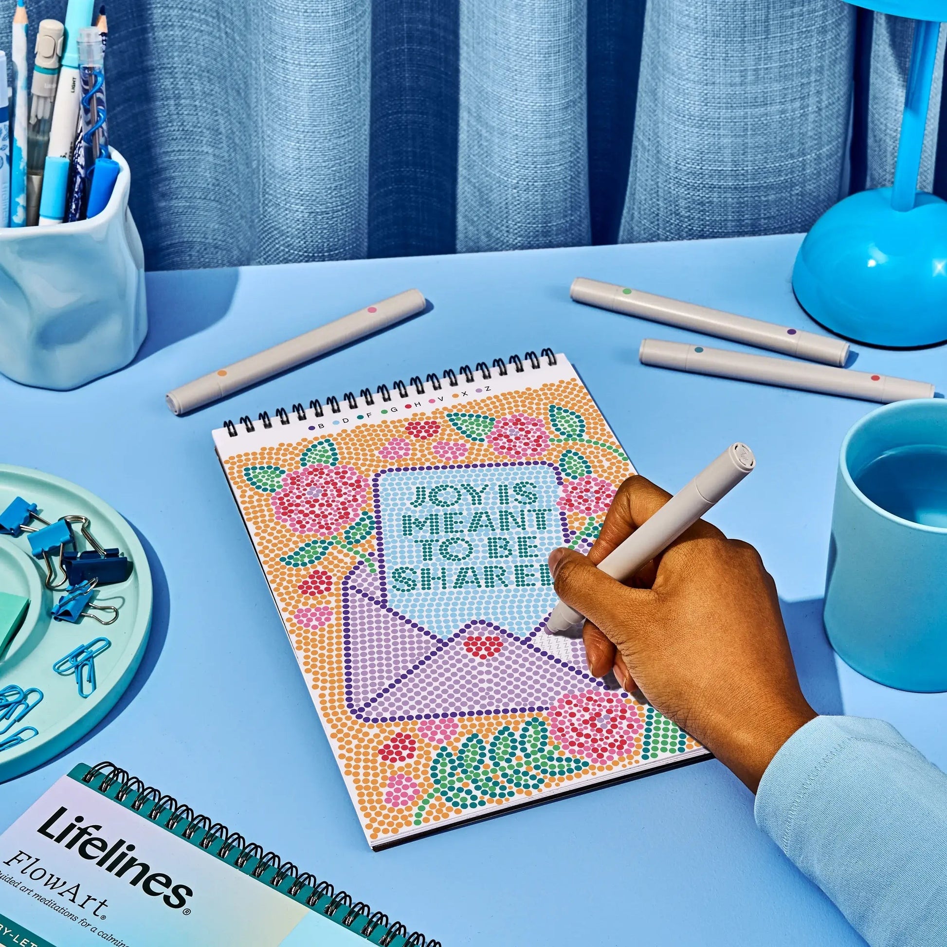 Mellow Monkey FlowArt dot-by-letter activity book with a colourful floral design on a blue desk with pens.