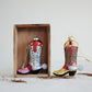 Mellow Monkey hand-painted glass cowboy boot ornaments with glitter and jewel accents in a rustic wooden crate