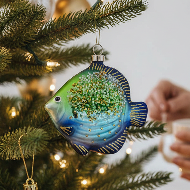Mellow Monkey glass fish ornament in blue and green with gold glitter and beads hanging on a decorated Christmas tree