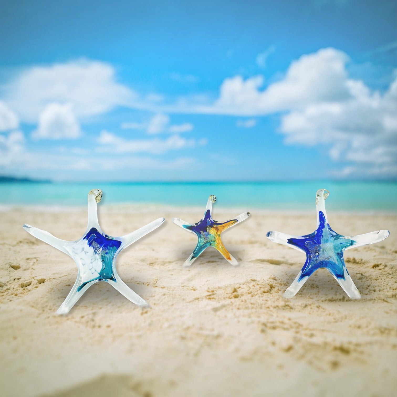 Mellow Monkey hand-blown glass star ornaments with blue and gold accents displayed on a sandy beach