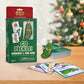 Mellow Monkey Go Pickle! ornament and card game set with a ceramic pickle ornament and playing cards on a wooden table with a blurred Christmas tree in the background.