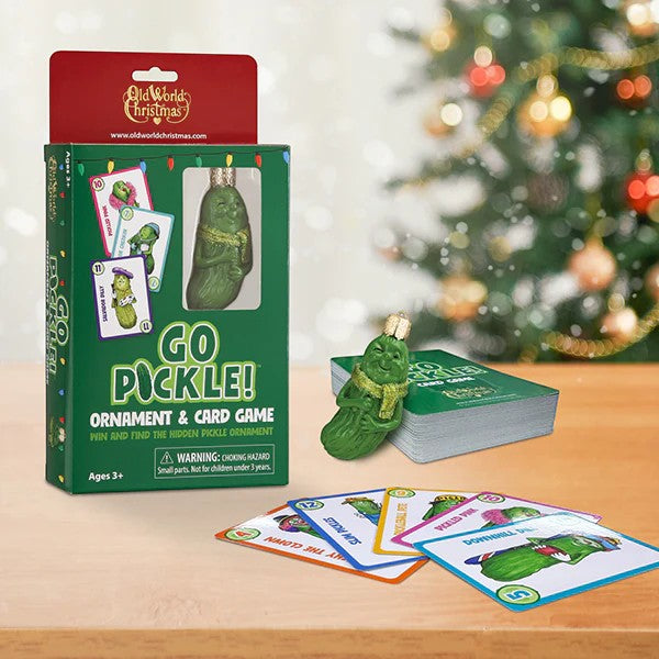 Mellow Monkey Go Pickle! ornament and card game set with a ceramic pickle ornament and playing cards on a wooden table with a blurred Christmas tree in the background.