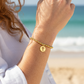 Mellow Monkey gold beaded stretch bracelet with a cut-out cross charm worn on a woman's wrist at the beach.