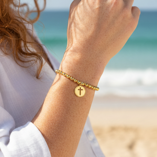 Mellow Monkey gold beaded stretch bracelet with a cut-out cross charm worn on a woman's wrist at the beach.