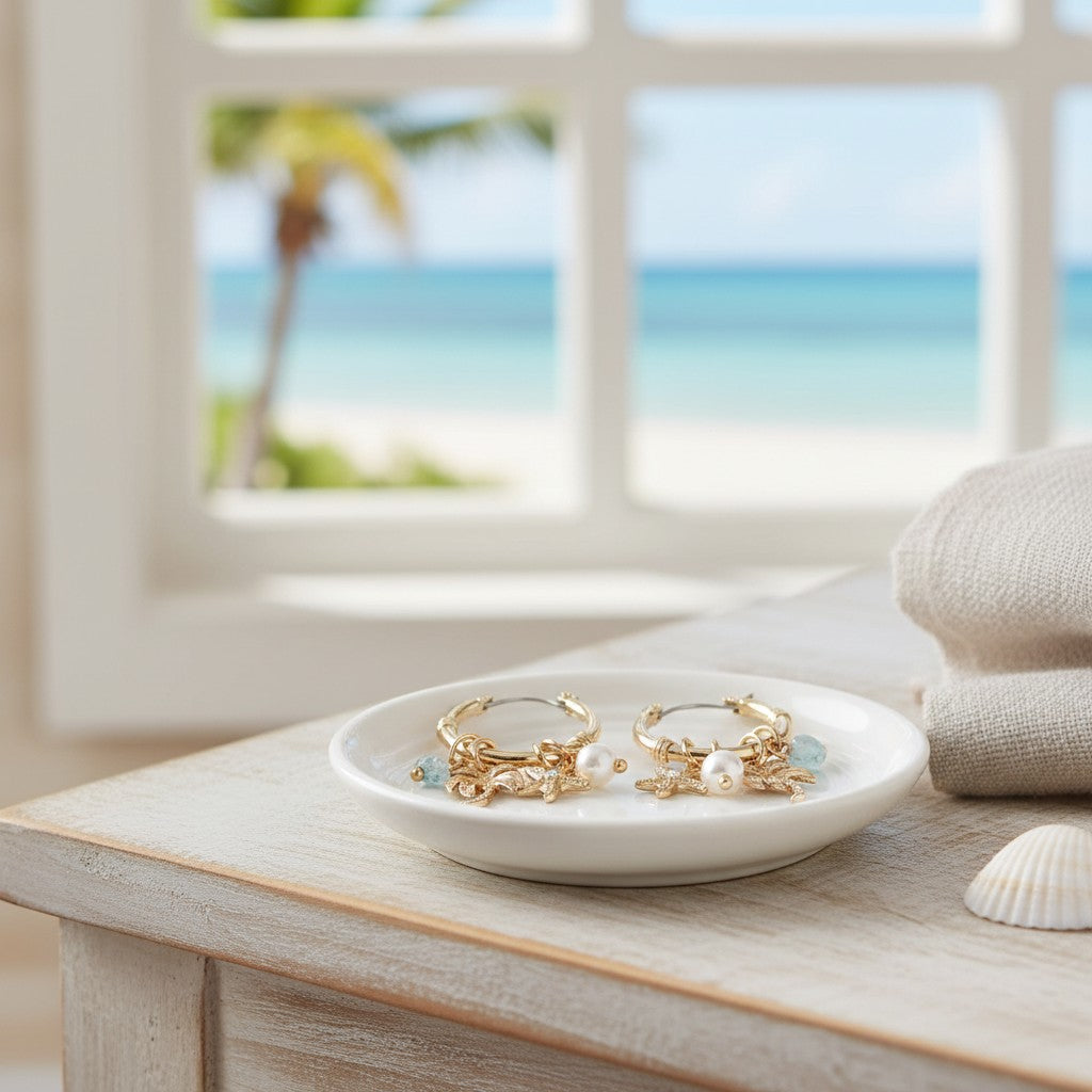 Mellow Monkey gold hoop earrings with coastal charms and pearls resting on a white dish by a beach view window.