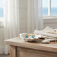 Mellow Monkey gold crab earrings featuring bright aqua crystals, displayed in a white shell dish on a wooden table.