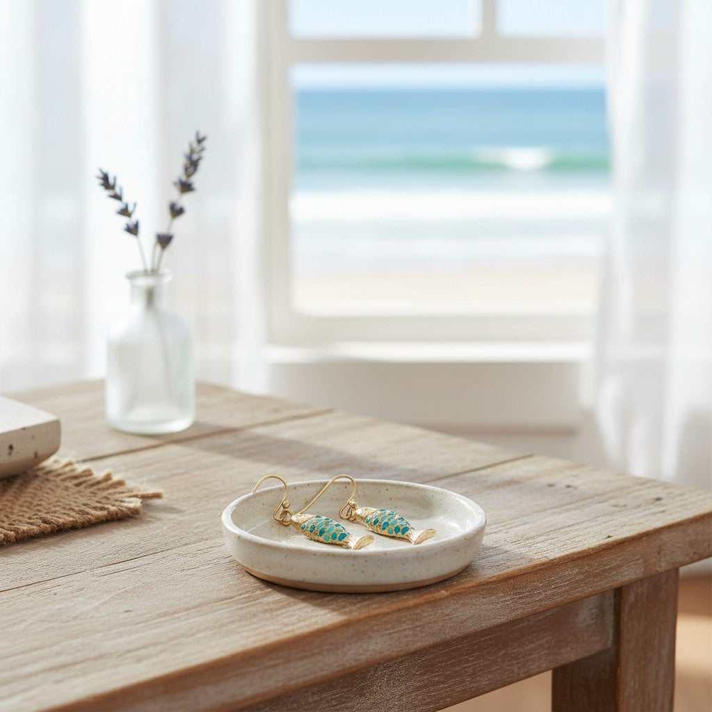 Mellow Monkey gold fish earrings with blue scales resting on a small ceramic dish by a beach window.