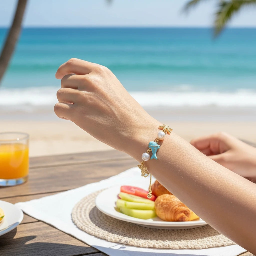 Mellow Monkey gold bracelet featuring a blue fish and starfish charms with pearls worn on a beachside table.