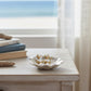 Mellow Monkey gold pearl drop earrings resting in a decorative shell dish on a wooden table by the beach.