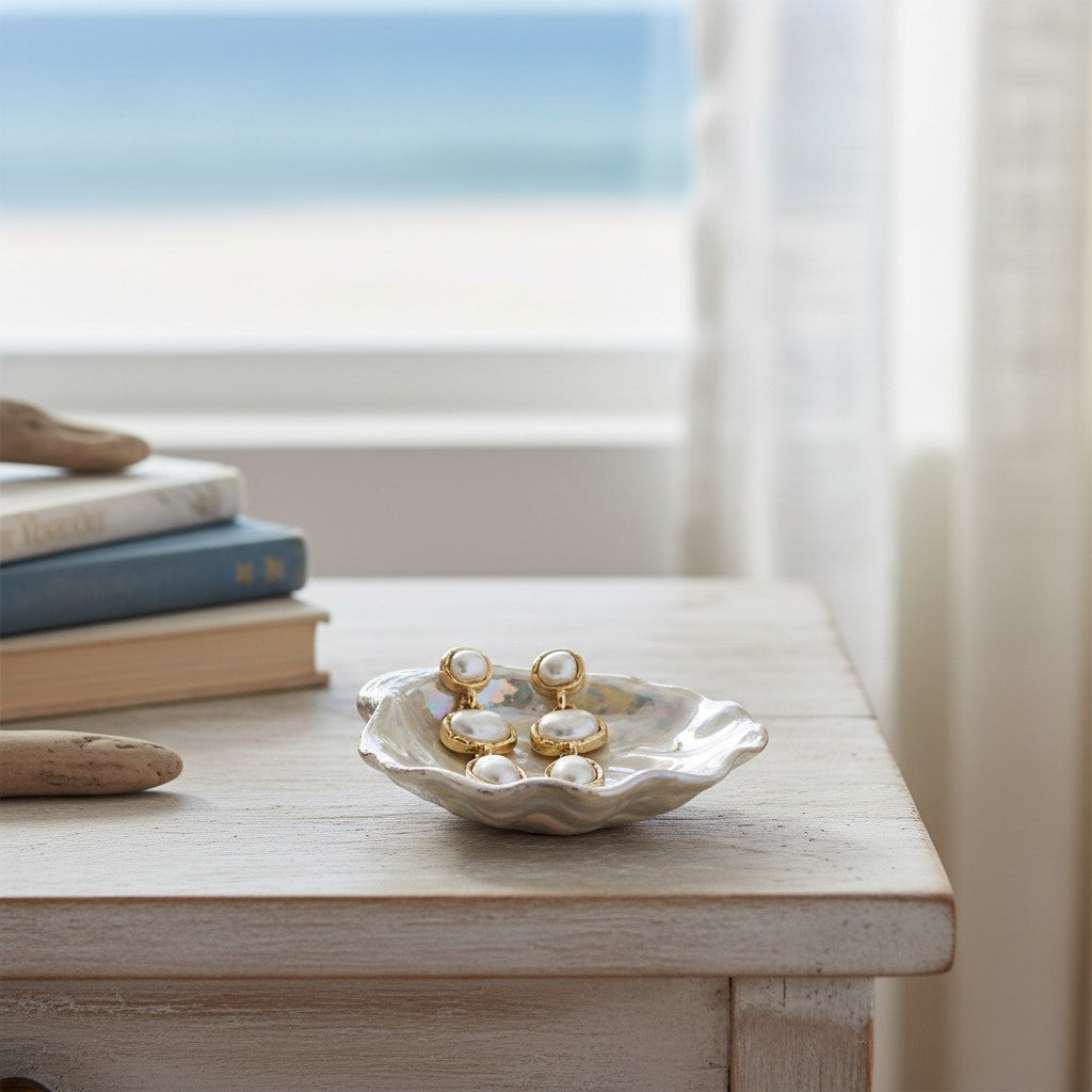 Mellow Monkey gold pearl drop earrings resting in a decorative shell dish on a wooden table by the beach.