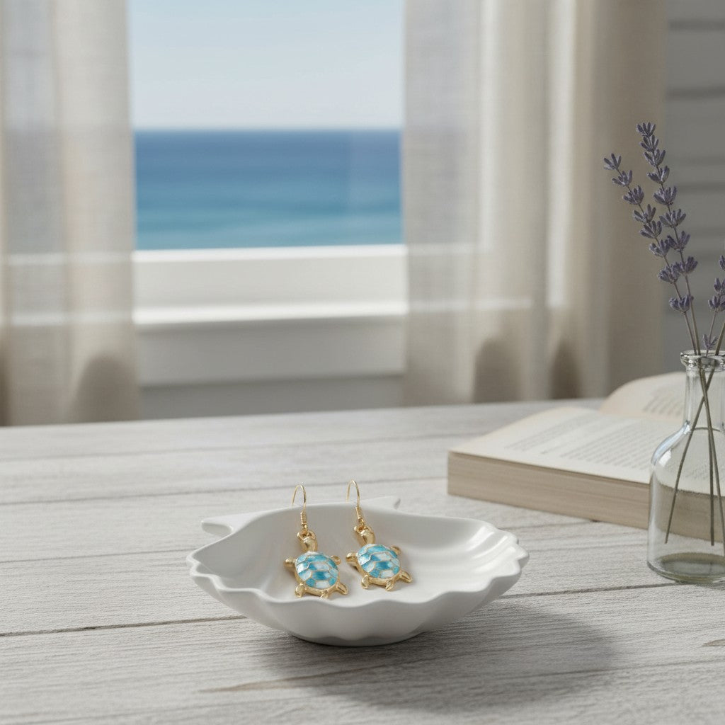 Mellow Monkey gold turtle earrings with blue marble enamel on a white shell dish by a coastal window.
