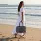 Mellow Monkey Grays Inlaid messenger bag in grey cotton and leather held by a woman in a white dress on the beach.