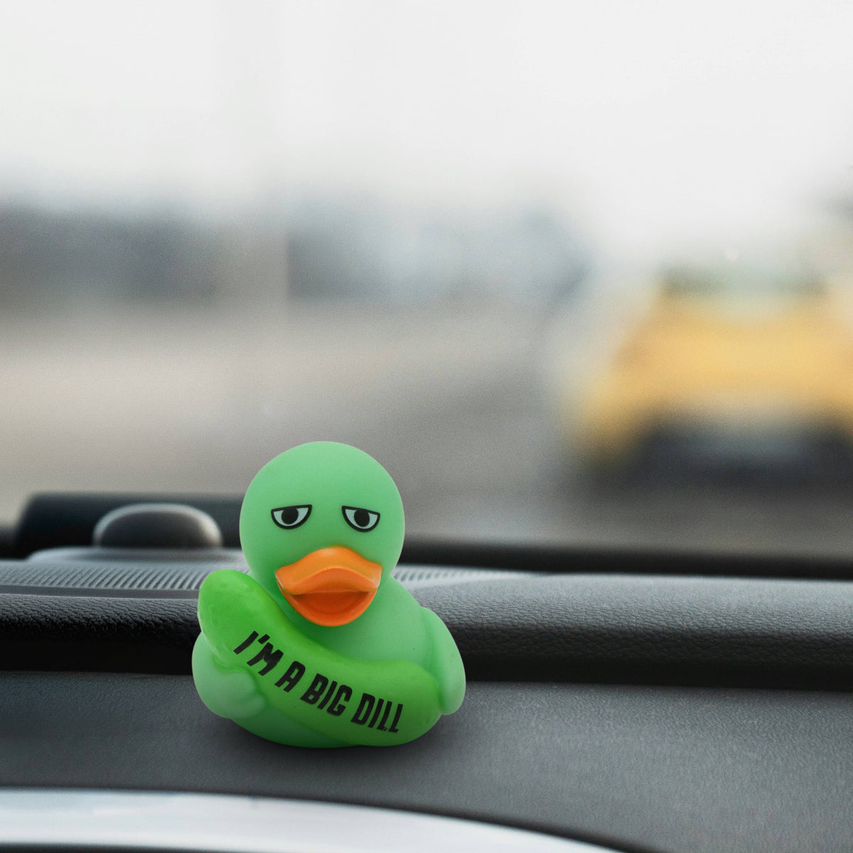 Mellow Monkey green rubber duck with an orange beak holding a pickle, displayed on a car dashboard.