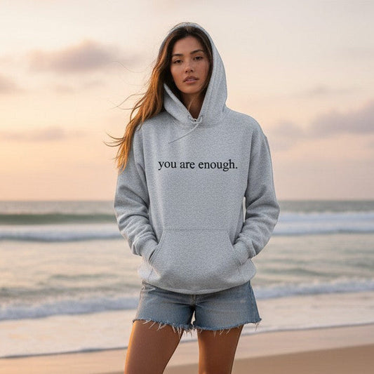 Mellow Monkey grey "You Are Enough" unisex hoodie with front pocket and drawstring hood, worn by a woman on a beach.