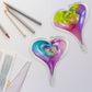 Two Mellow Monkey hand-blown glass heart ornaments with vibrant multicolored swirls and glittery accents on a desk.