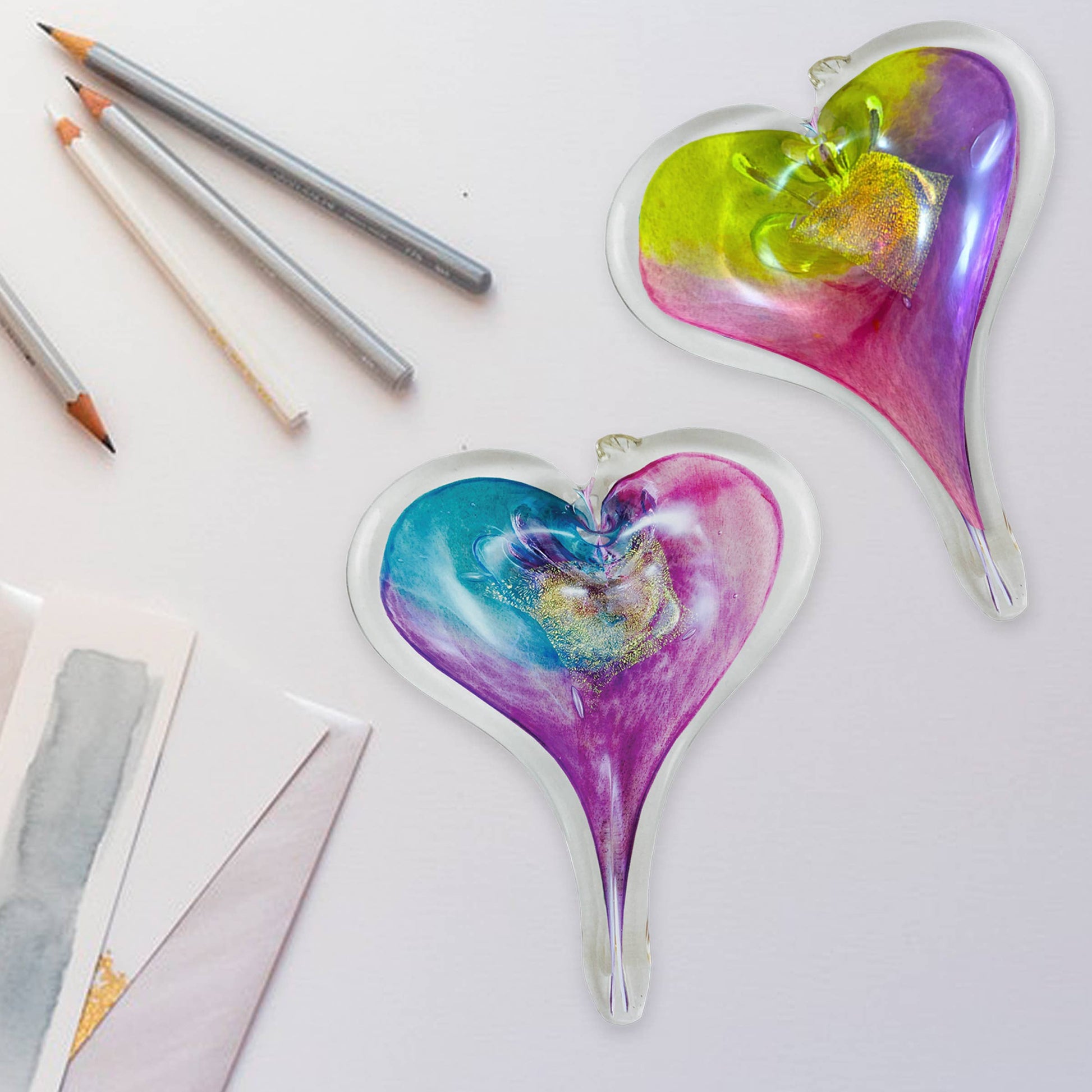 Two Mellow Monkey hand-blown glass heart ornaments with vibrant multicolored swirls and glittery accents on a desk.