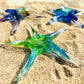 Mellow Monkey hand-blown glass star ornaments in blue and green resting on sandy beach surface