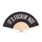 Mellow Monkey black cotton hand fan with white text reading 'IT'S F*CKIN' HOT' and wooden ribs.
