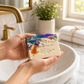 Mellow Monkey Deck The Halls handmade bar soap with multicolored holiday light swirls being rinsed under a faucet.