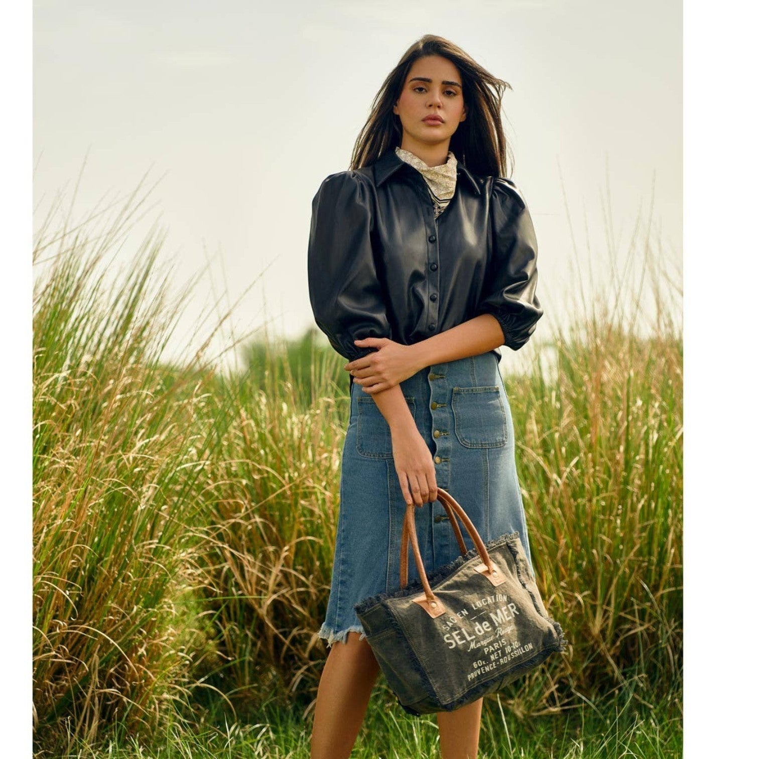 Model holding a Mellow Monkey Hazy Tones canvas and leather crossbody bag with vintage typography in a field.