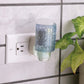 Mellow Monkey Hobnail WallScents fragrance warmer in blue glass plugged into a wall outlet next to a green plant.