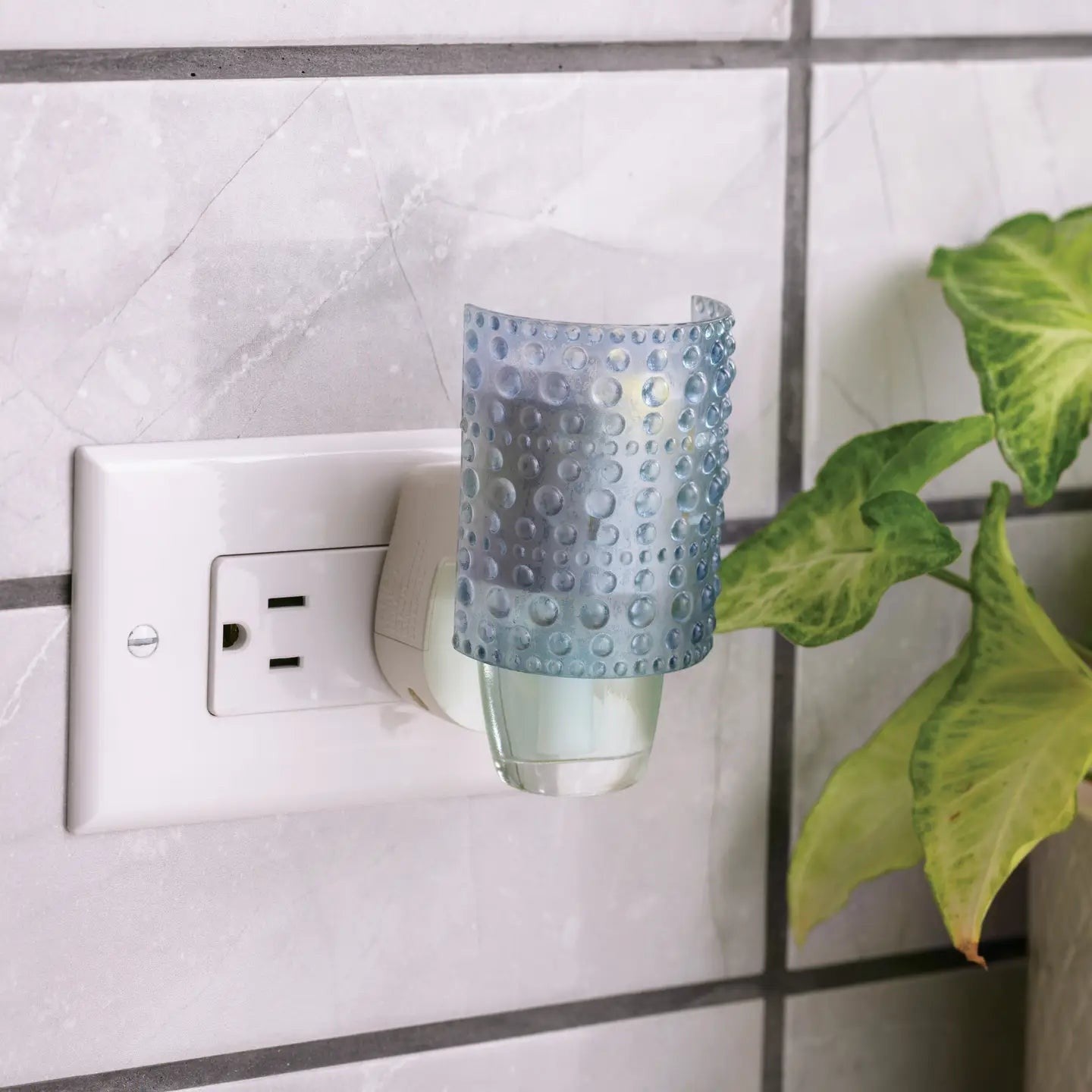 Mellow Monkey Hobnail WallScents fragrance warmer in blue glass plugged into a wall outlet next to a green plant.