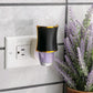 Mellow Monkey Hourglass WallScents fragrance warmer in black with gold trim plugged into a wall outlet.