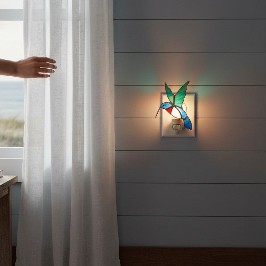 Mellow Monkey stained glass hummingbird nightlight glowing on a wall next to a window with sheer curtains
