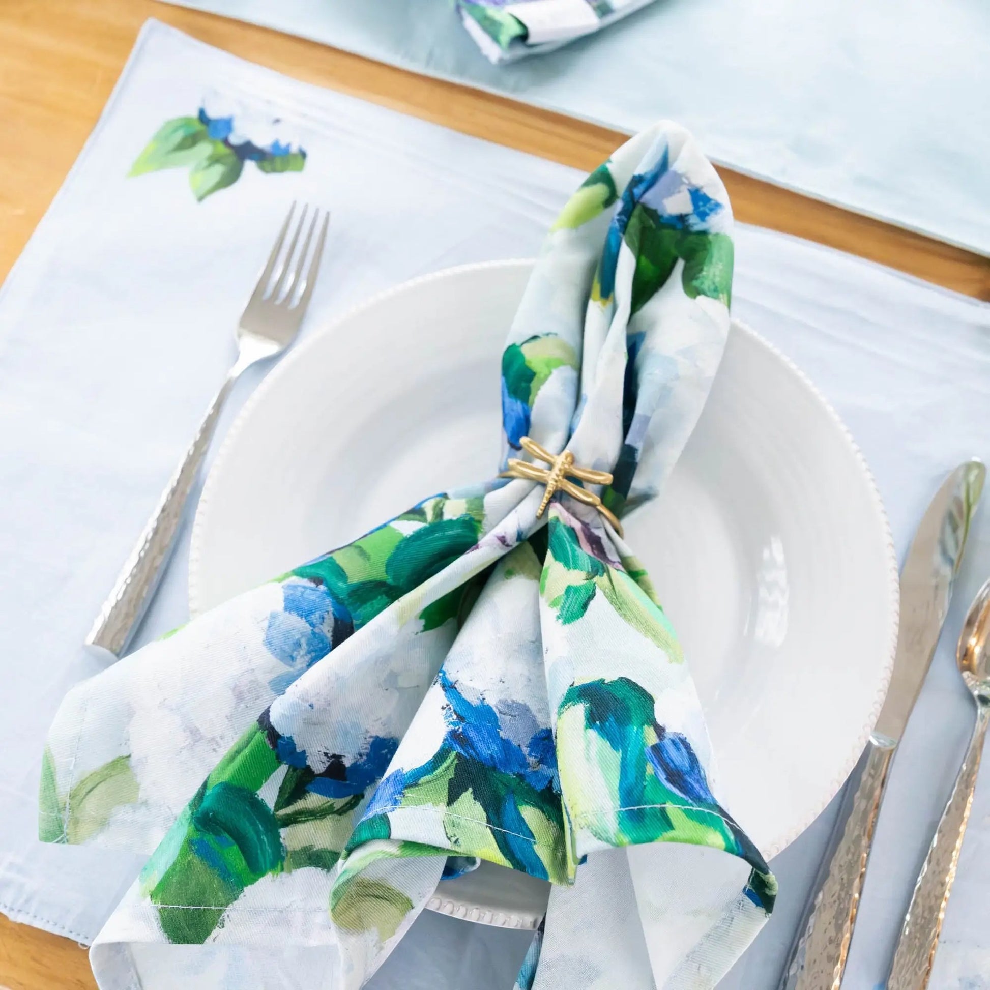 Mellow Monkey cotton dinner napkin with blue and green hydrangea floral print, folded on a white plate with a ring.