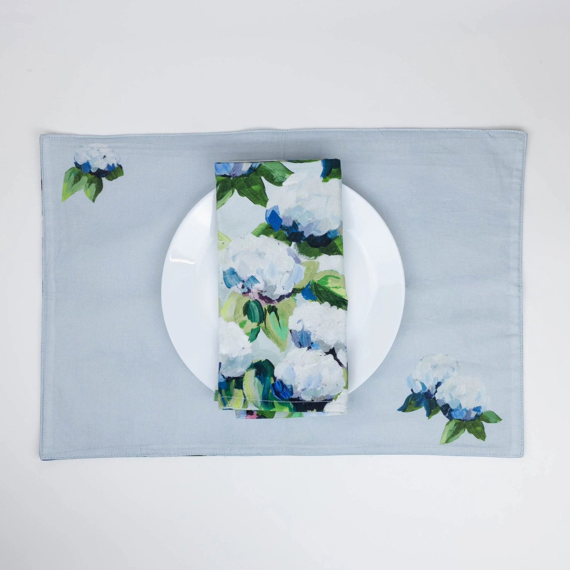 Mellow Monkey reversible woven cotton placemat featuring blue and white hydrangea artwork on a light blue background.
