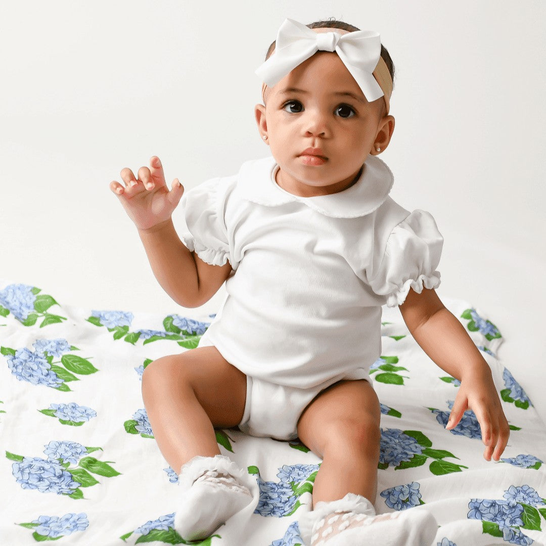 Mellow Monkey baby sitting on a soft white muslin swaddle blanket featuring a blue hydrangea floral print