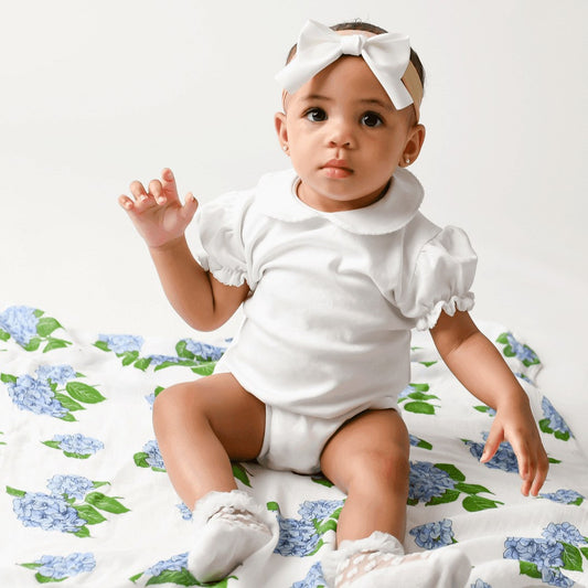 Mellow Monkey baby sitting on a soft white muslin swaddle blanket featuring a blue hydrangea floral print