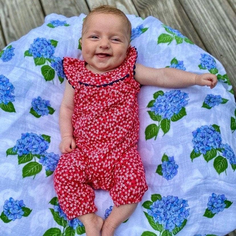 Mellow Monkey hydrangea print muslin swaddle blanket with a smiling baby in a red floral romper lying on top