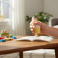 Mellow Monkey 3-inch glittery ice cream squeeze toy being held by a hand over a wooden desk with a book and blocks.