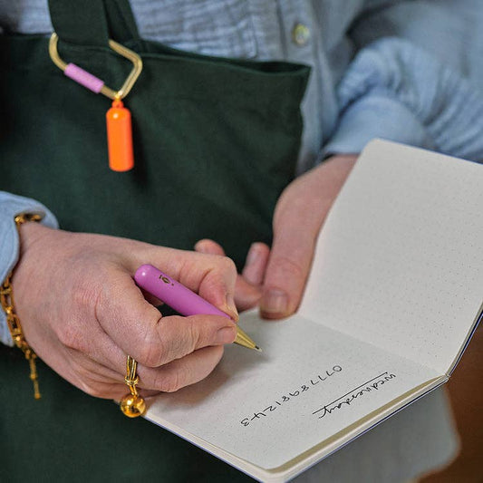 Mellow Monkey Inkerie pocket-sized purple ballpoint pen with a bright orange carabiner being used in a notebook