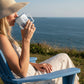 Mellow Monkey 12oz insulated stainless steel mug with art, held by a woman in a sun hat sitting by the ocean.