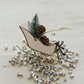 Mellow Monkey white and gold iron sleigh ornament featuring a bottle brush tree, pinecones, and a green bow.