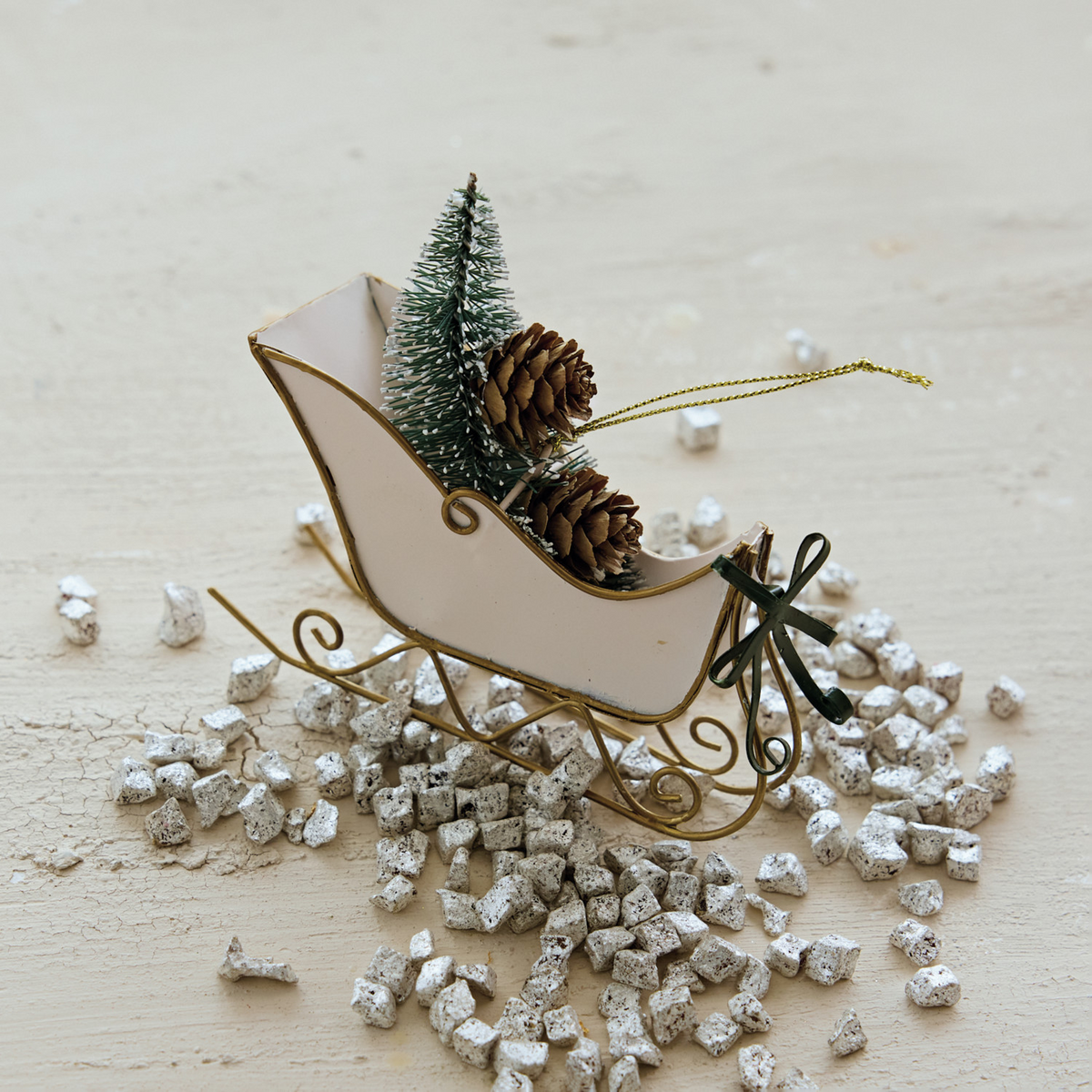 Mellow Monkey white and gold iron sleigh ornament featuring a bottle brush tree, pinecones, and a green bow.