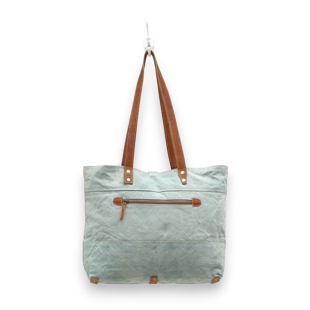 Mellow Monkey La Couspaude canvas tote bag in light blue with brown leather handles and a zippered exterior pocket.