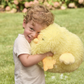 Mellow Monkey large yellow Snuggle Puff duckie plush toy being hugged by a smiling young boy in a grassy garden.
