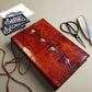 Mellow Monkey reddish-brown leather journal with embossed elemental symbols and gemstone accents, cord closure.