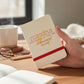 Mellow Monkey leatherette pocket journal with red embossed text and elastic band held in a hand on a wooden desk
