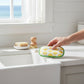 Mellow Monkey absorbent kitchen sponge with yellow lemon pattern and green trim held over a white farmhouse sink.