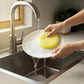 Mellow Monkey Citrus Squeeze Scrubby washing a white plate under a kitchen faucet with yellow lemon-shaped design