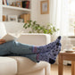 Mellow Monkey light blue crew socks with dog graphics and text worn by a person relaxing on a sofa with a book.