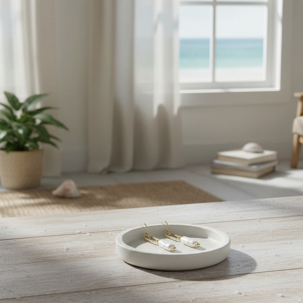 Mellow Monkey linear pearl and gold earrings resting in a white ceramic dish on a wooden table with a beach view.