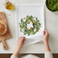 Mellow Monkey white cotton flour sack towel featuring a watercolor magnolia wreath design on a kitchen counter.